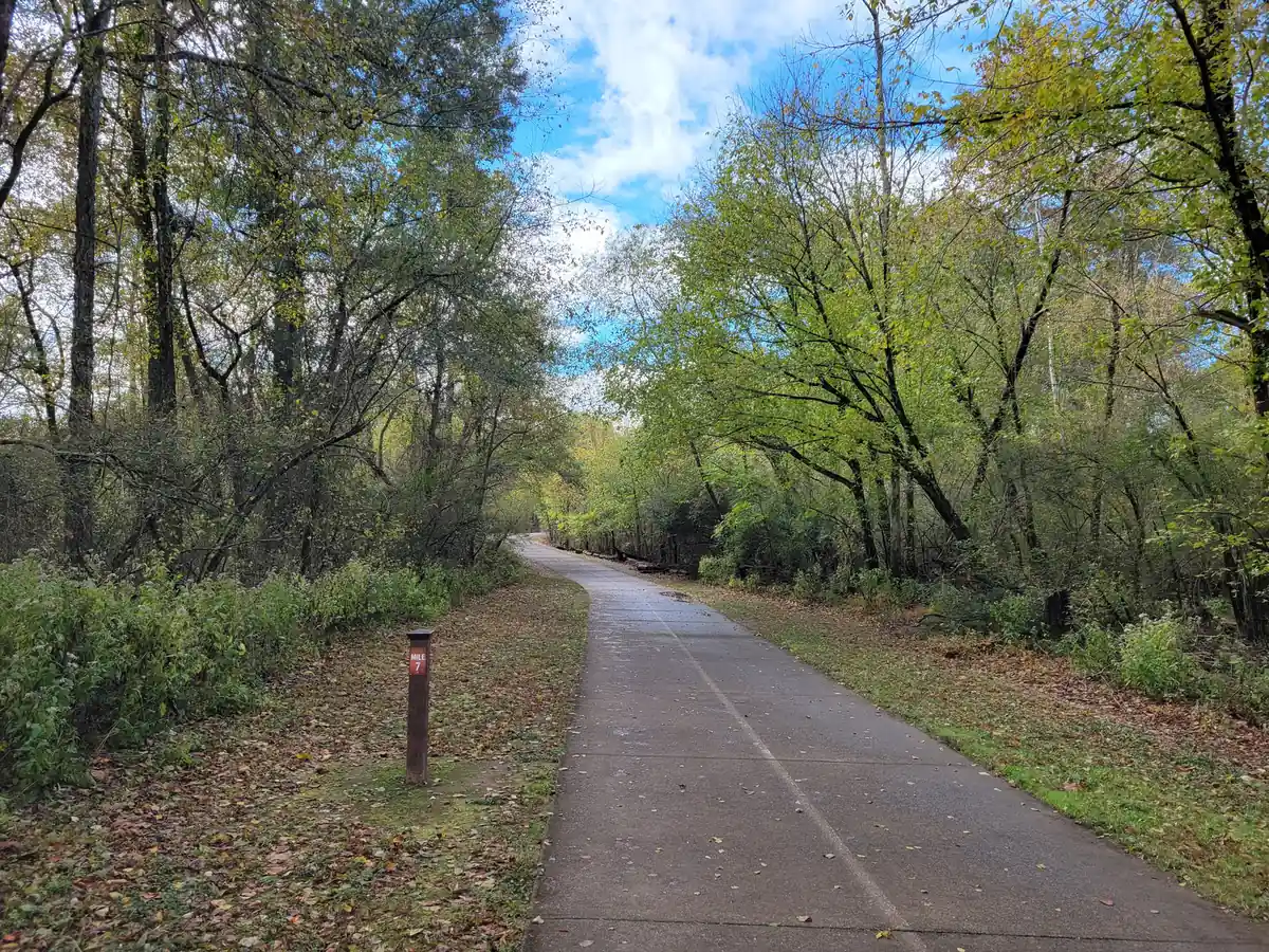 A concrete multi-use path with a faint center line curves gently into the distance between autumn trees with yellowing and green leaves, with fallen leaves on the grassy verges, a small brown mile-marker post at left and blue sky with white clouds overhead.