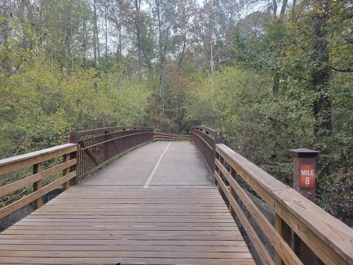 A wooden boardwalk bridge with brown metal and timber railings and a painted center line leads straight ahead into dense autumn woodland; a brown post with an orange 'Mile 8' plate stands at the right railing.