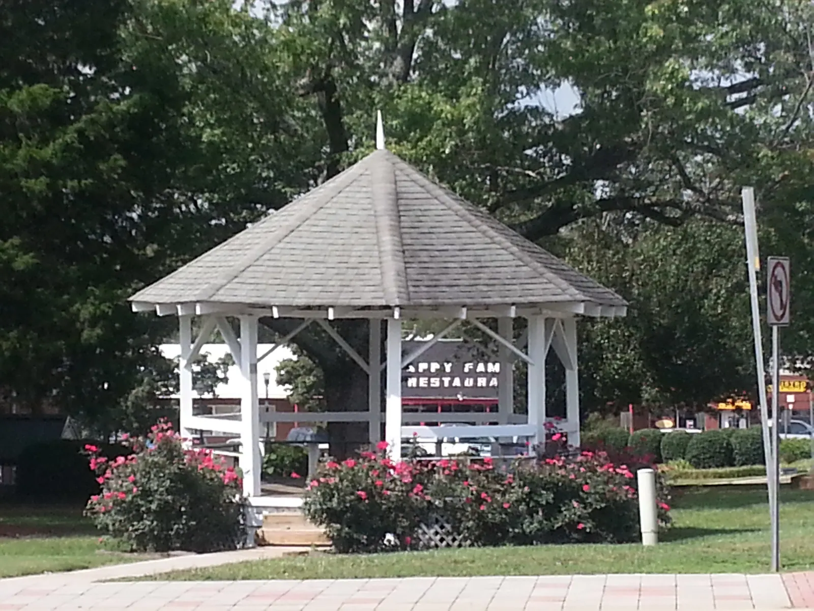 A white octagonal wooden gazebo with a grey shingled roof and lattice skirting sits on a lawn ringed by pink rose bushes, with large shade trees behind, a 'No left turn' road sign on a pole at right and a partially hidden restaurant sign in the background.