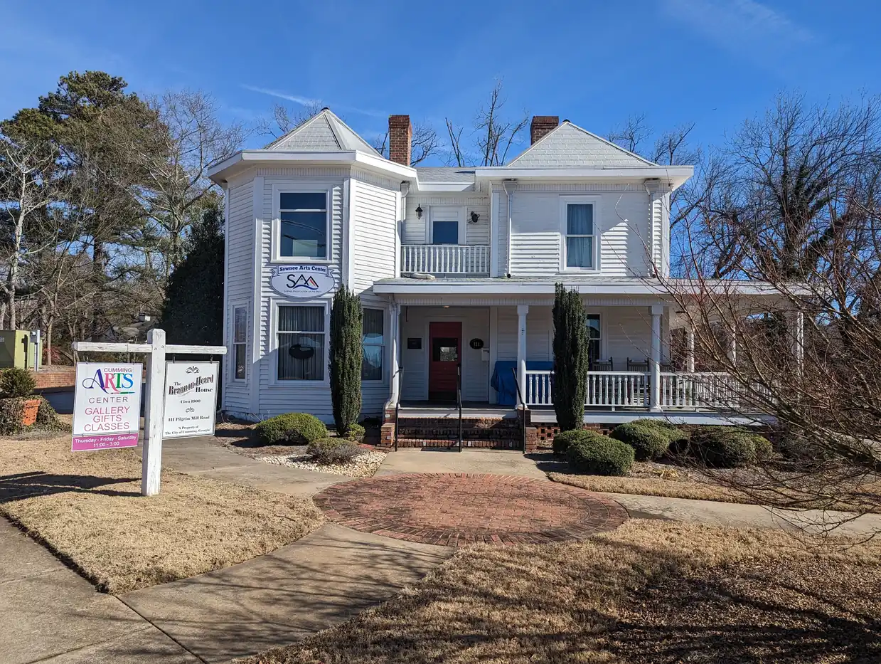 A white two-story Victorian-style house with a corner bay, a wraparound porch with white railings, a red front door and two brick chimneys stands among bare winter trees; a yard sign reads 'Cumming Arts Center – Gallery, Gifts, Classes' beside a second sign naming the house, and an oval sign with the letters 'SAA' hangs on the facade.