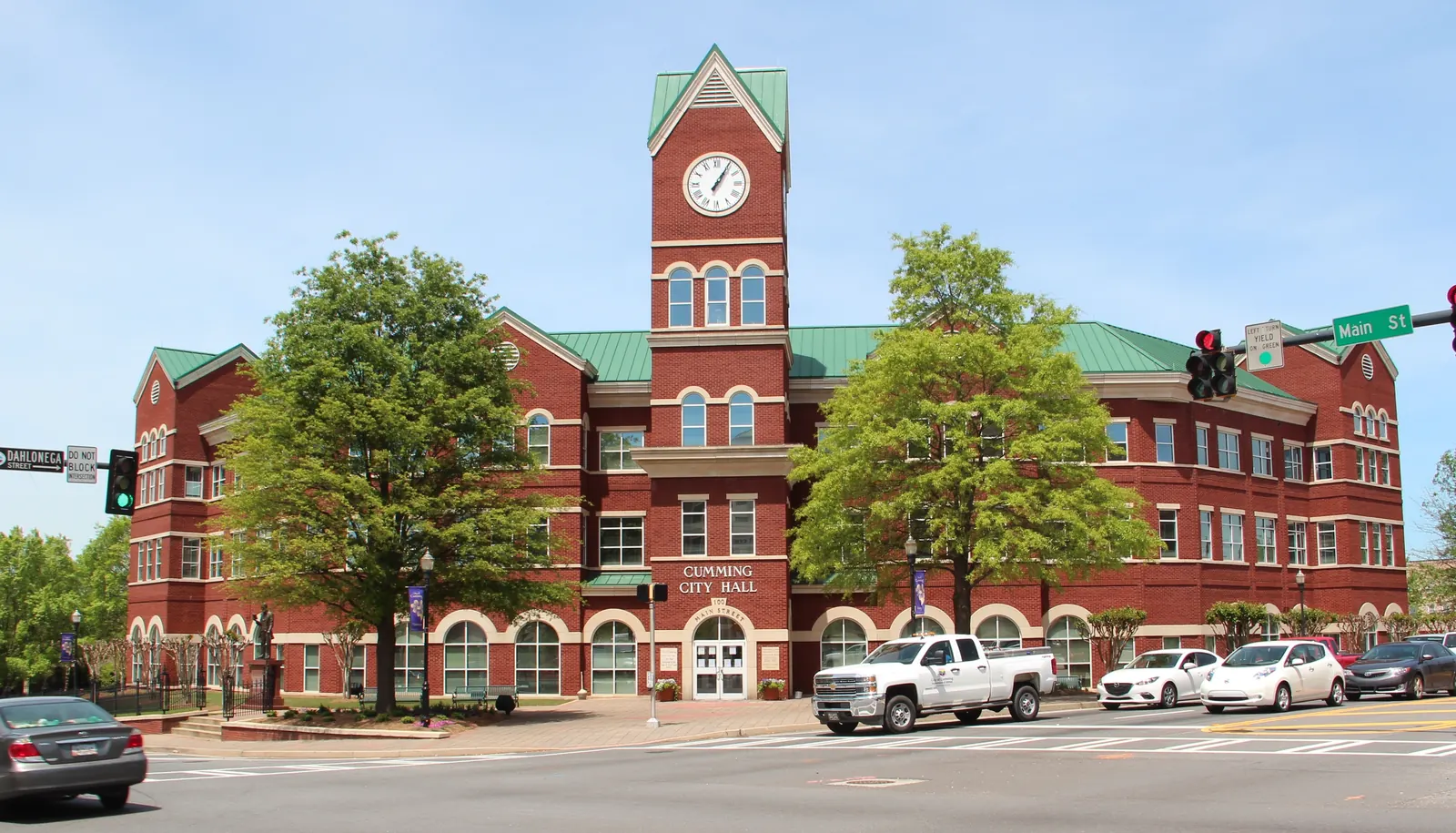 A three-story red-brick building with a green metal roof, arched ground-floor windows and a central clock tower stands behind a street intersection under a blue sky; 'Cumming City Hall' lettering is above the entrance, two trees frame the tower, pickup trucks and cars are parked in front, and a traffic signal with a 'Main St' street sign hangs at right.