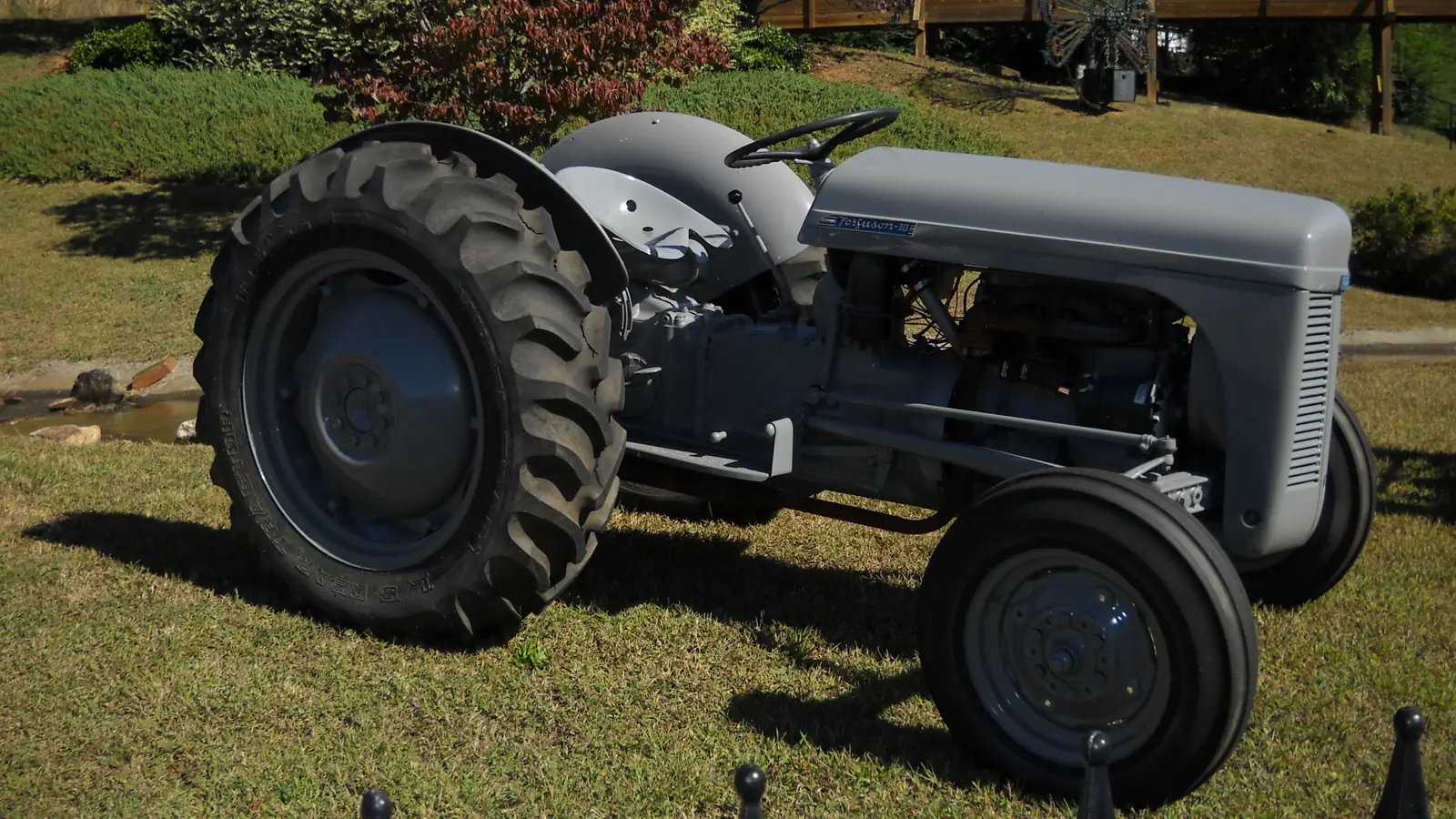 A restored grey vintage farm tractor with a 'Ferguson' badge on the hood, large ribbed rear tires and grey-painted wheels stands on a mown lawn in bright sun, with landscaped shrubs, a small stream bed and a wooden deck behind and black post tops along the bottom edge.