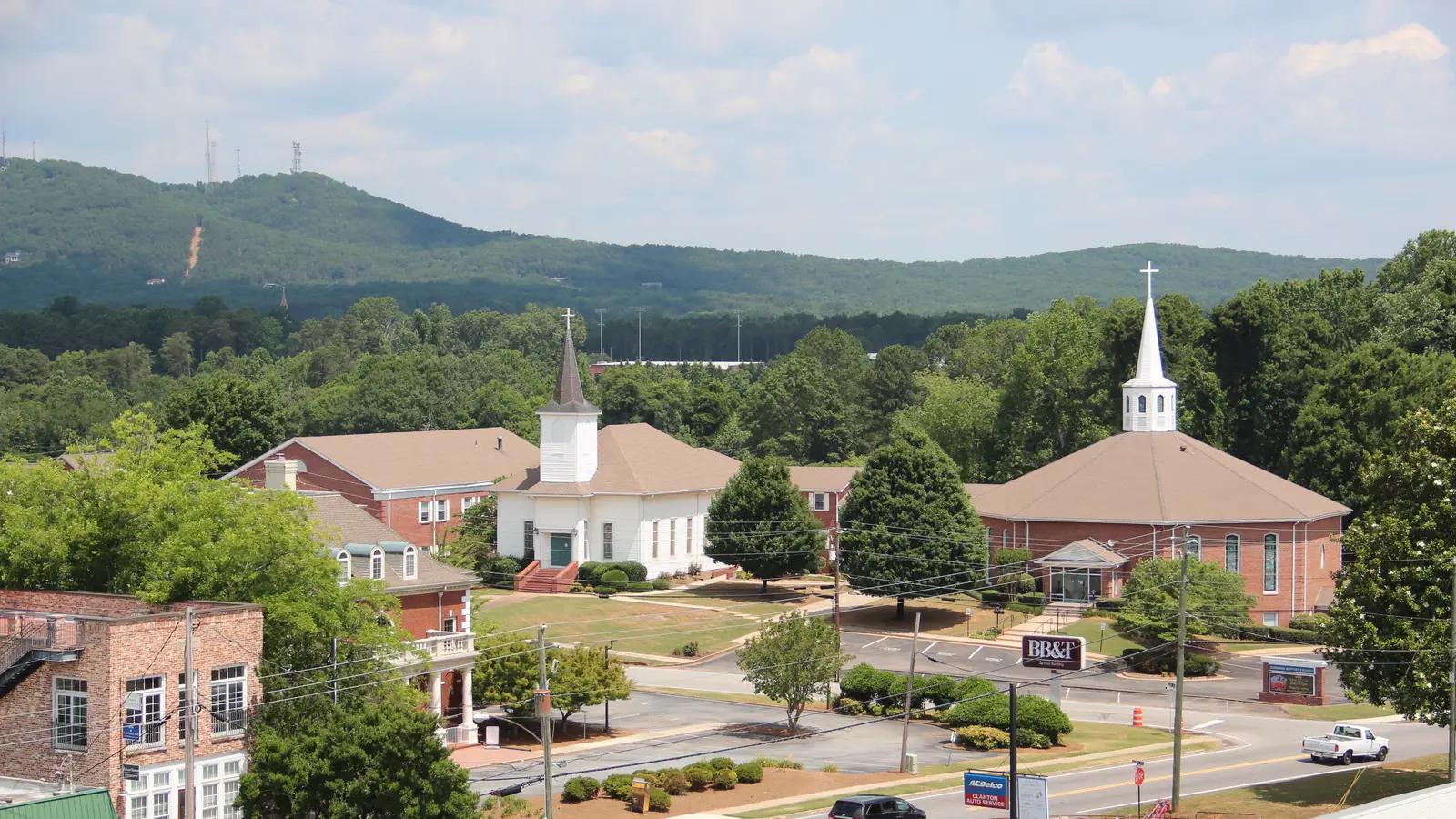 Elevated view of a white-steepled church and a red-brick church with a white steeple set among lawns, trees and a parking lot along a road with cars, with older brick commercial buildings at left and a forested mountain ridge with towers and a cleared slope line across the horizon.