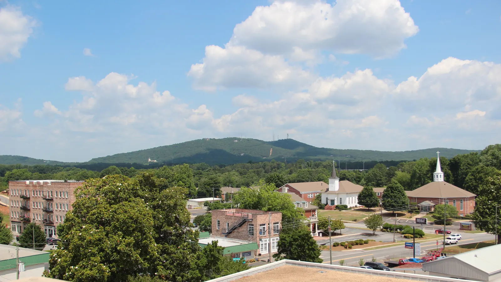 From a flat rooftop, a view over downtown rooftops, a multi-story brick building, two white church steeples and tree-lined streets toward a long, forested mountain ridge topped by communication towers, under a blue sky with large white cumulus clouds; a beige parapet and roof surface fill the bottom edge.