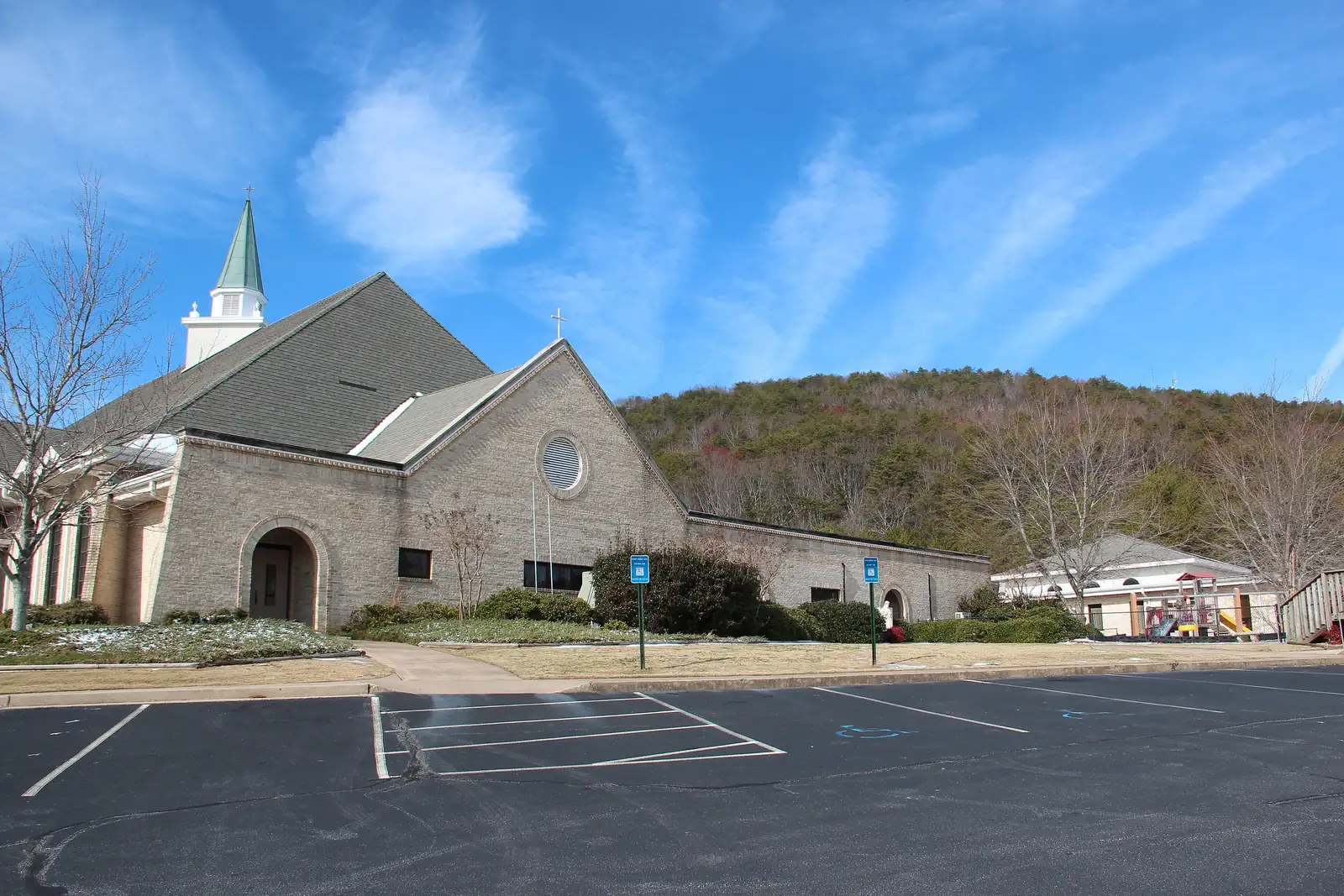 A tan brick church with a steep grey roof, a round window and a white steeple with a green spire sits behind an empty parking lot with patches of snow, with a wooded hillside of bare winter trees rising behind it under a blue sky streaked with wispy clouds.