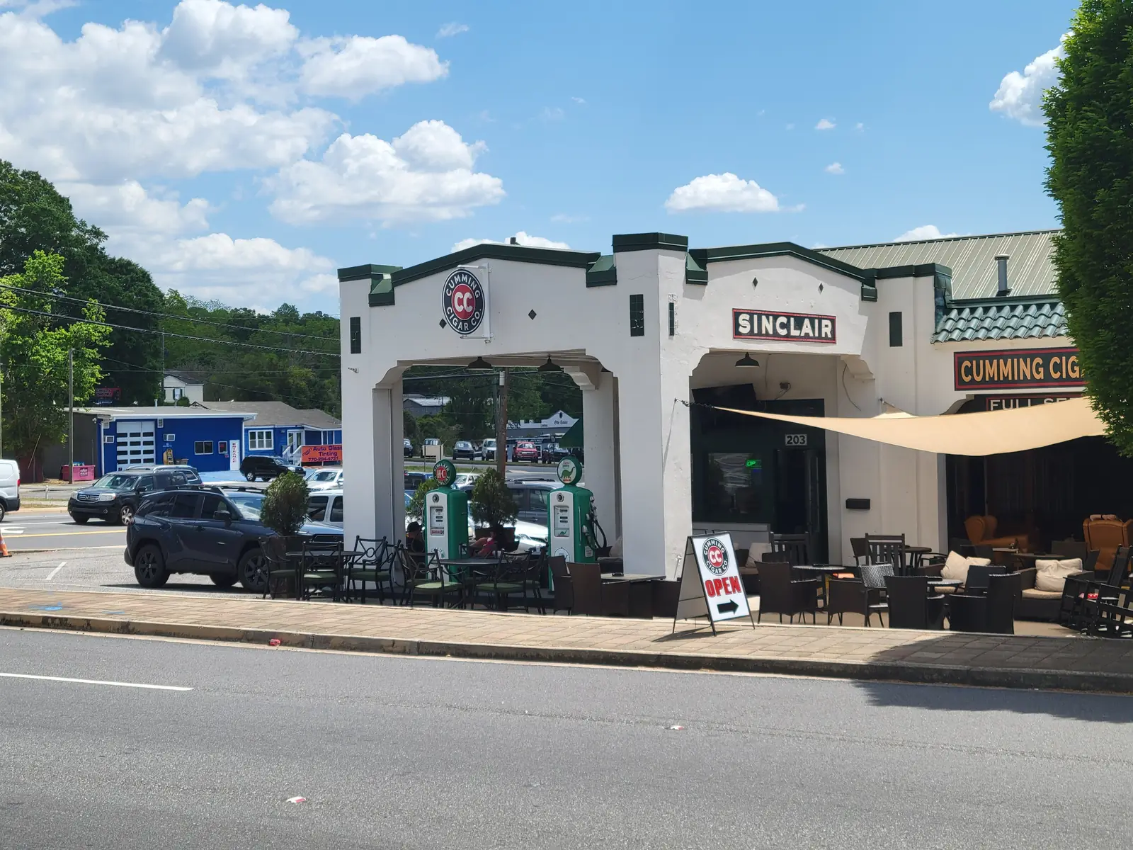 A white stucco former gas station with dark green trim, an open canopy bay, two vintage-style green fuel pumps and a 'Sinclair' sign now operates as a cafe/lounge with patio tables, chairs and a sidewalk 'Open' sign; a sign reading 'Cumming Cig…' is partly visible at right, cars are parked to the left and wooded hills rise under a blue sky with puffy clouds.