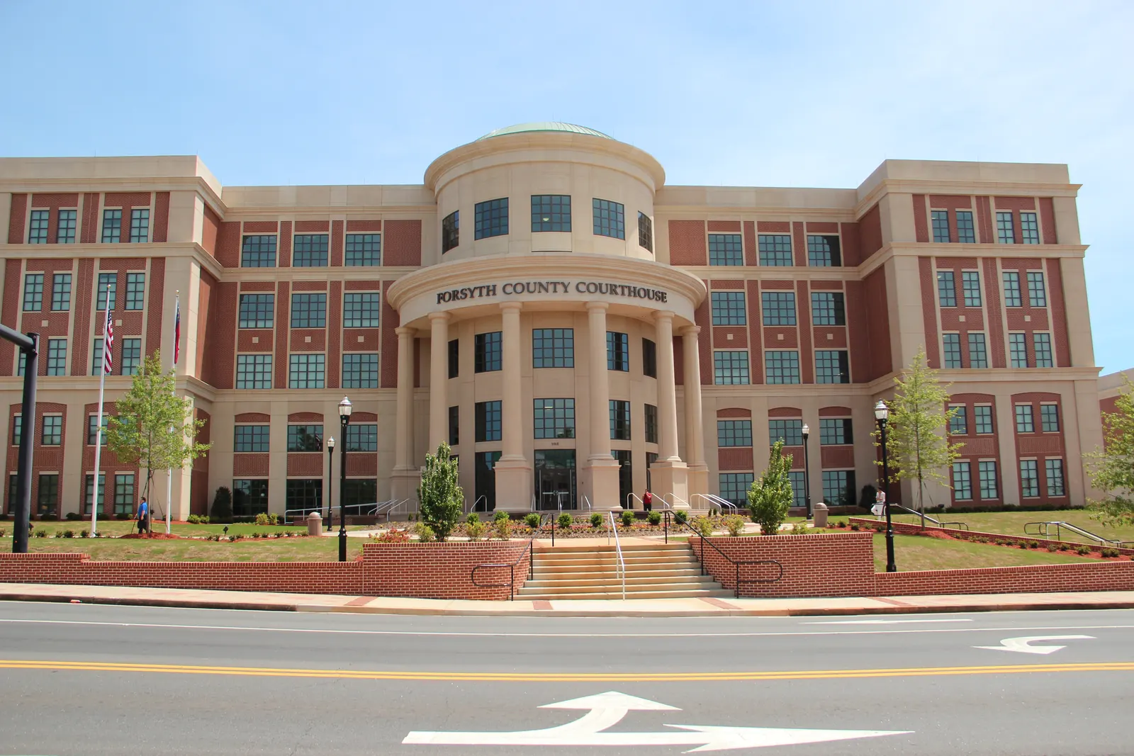 A large four-story building of red brick and cream stone with a rounded, columned entrance rotunda lettered 'Forsyth County Courthouse' rises behind brick retaining walls, steps and young trees, seen across an empty two-lane street with white turn arrows under a pale blue sky.