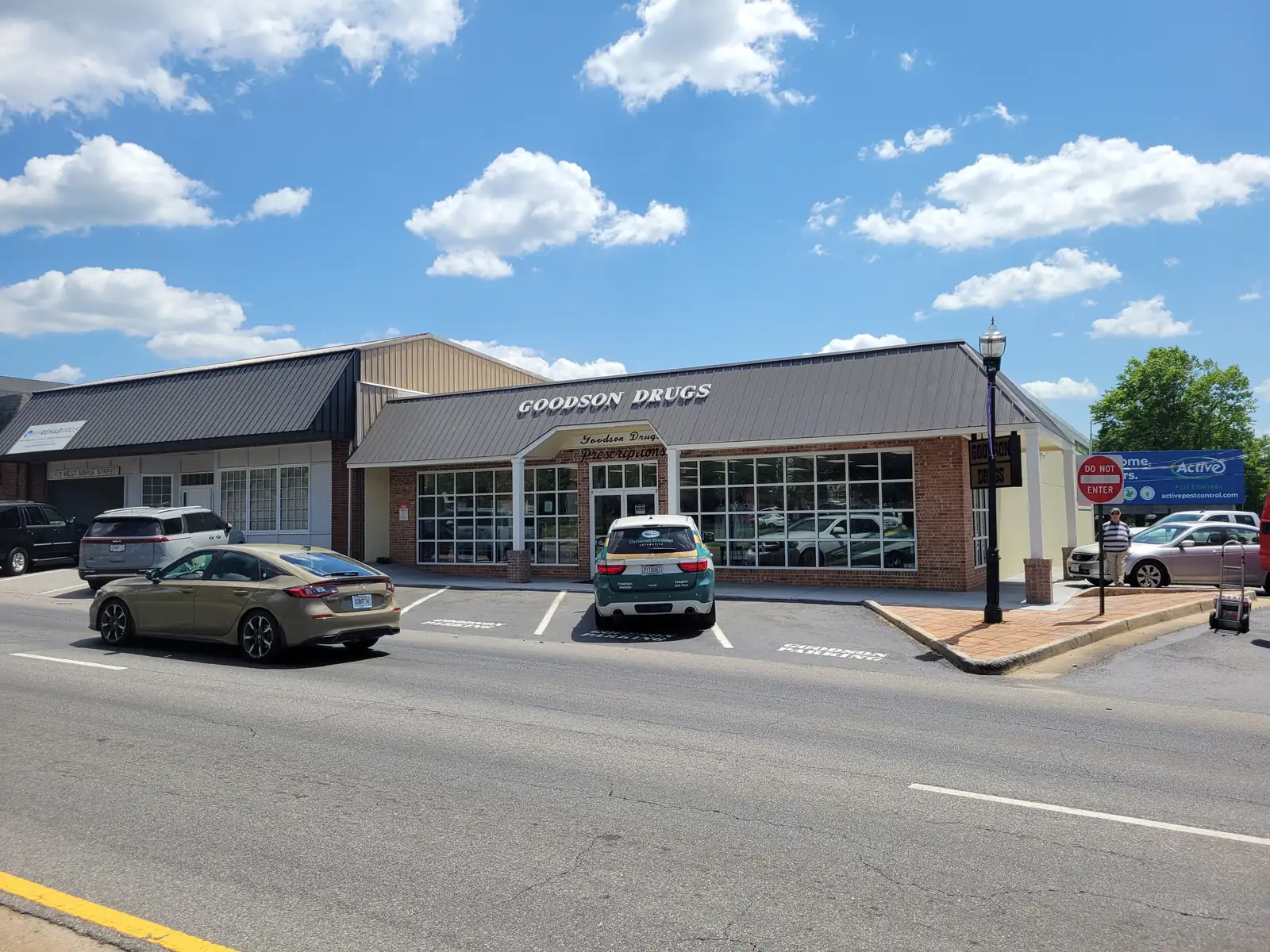 A low brick-and-glass storefront with a grey metal mansard roof lettered 'Goodson Drugs' sits beside an older white-roofed commercial building, with cars parked in angled spaces in front, a black street lamp, a 'Do Not Enter' sign and a blue banner at right, under a blue sky with scattered white clouds.