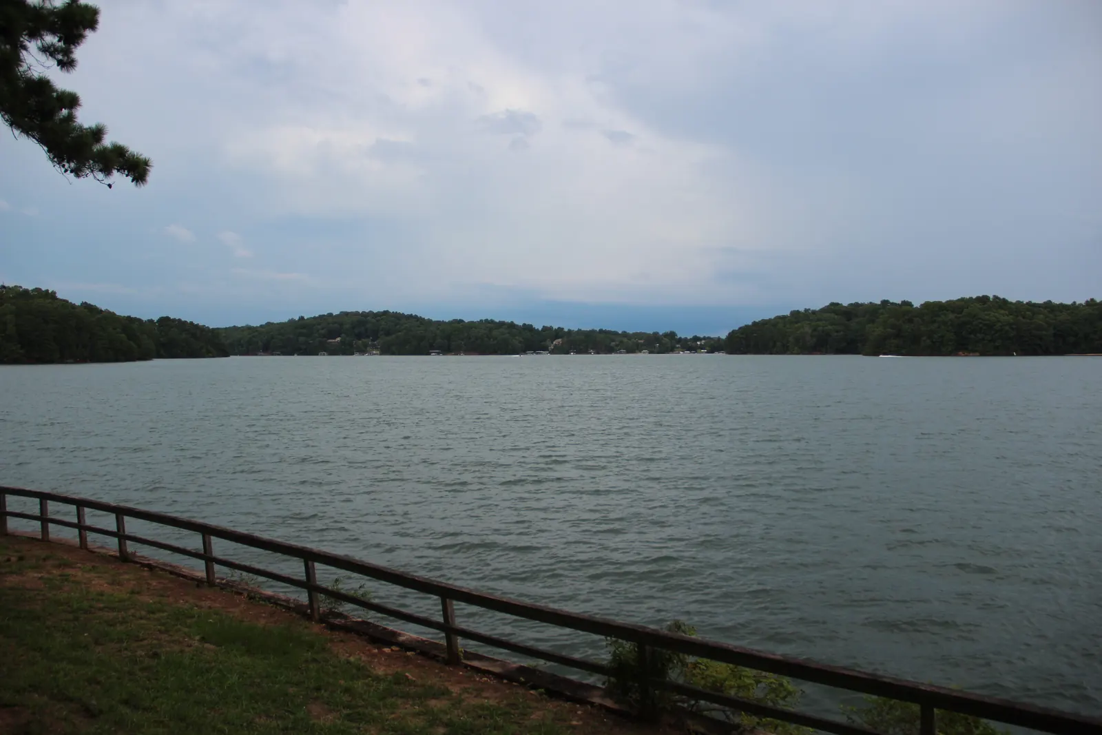 A wide stretch of grey-green lake water ruffled by wind extends to low forested shorelines under a heavy, cloudy sky; a wooden rail fence runs along a grassy bank in the foreground and a pine bough hangs in the upper-left corner.