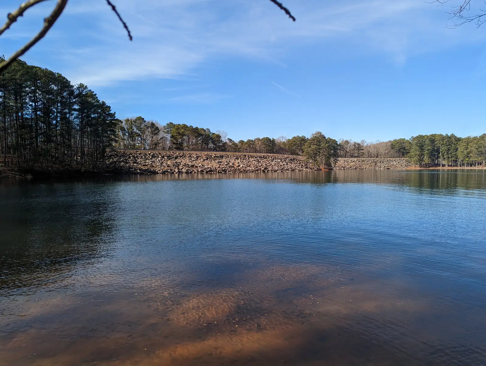 Calm blue lake water in the foreground, clear enough to show the sandy-red bottom, reflects a long rock-faced embankment topped with trees and a stand of tall pines at left under a bright blue sky with thin clouds; bare twigs dip into the top edge of the frame.
