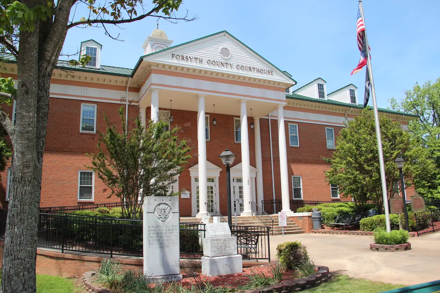 A two-story red-brick building with a white columned portico, a pediment lettered 'Forsyth County Courthouse', dormer windows and a small white cupola stands behind a granite memorial stone, an iron fence and landscaped beds; an American flag and a Georgia flag fly on a tall pole at right, and a tree trunk frames the left edge.