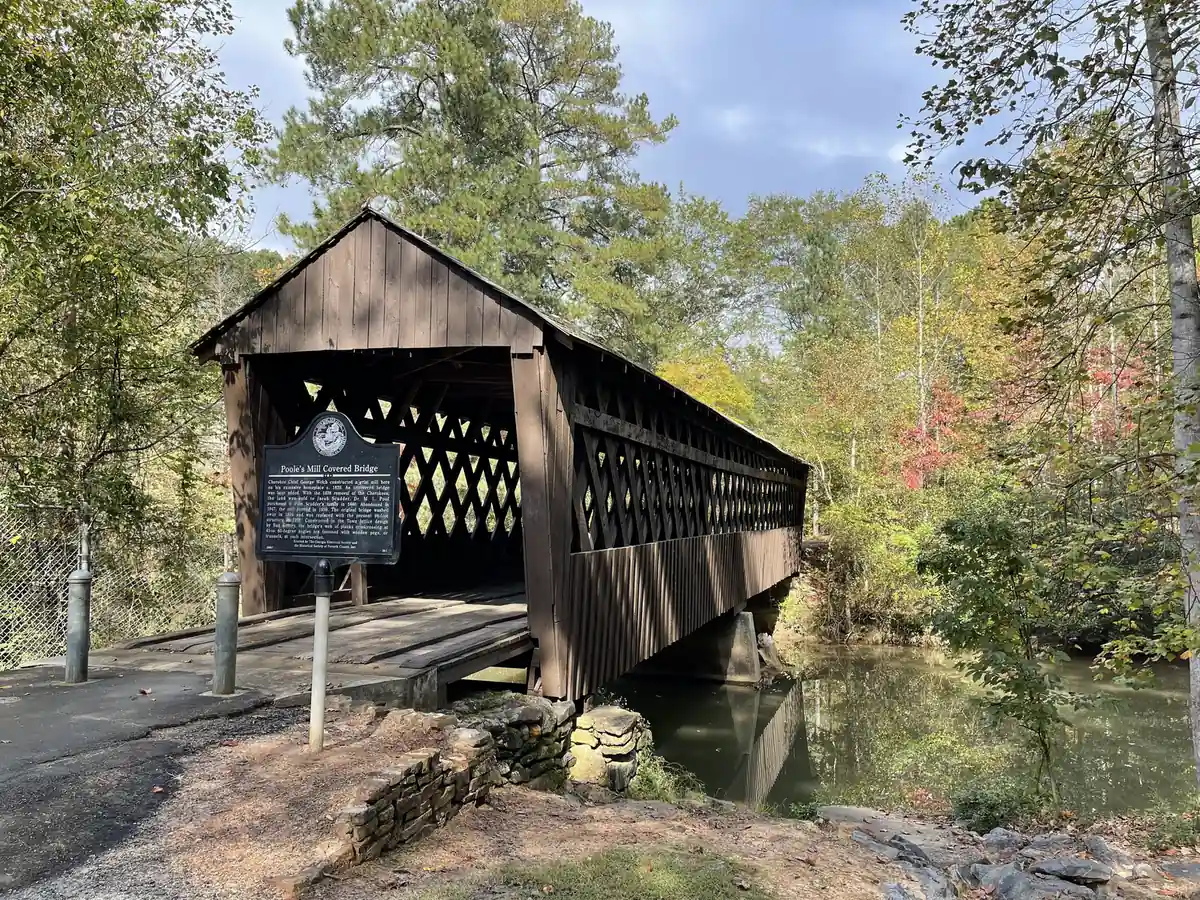 A dark-brown wooden covered bridge with lattice-truss sides spans a calm creek among early-autumn trees, with a black Georgia historical marker titled 'Poole's Mill Covered Bridge' on a post in the foreground, a stone wall and paved path at left and a chain-link fence at far left.