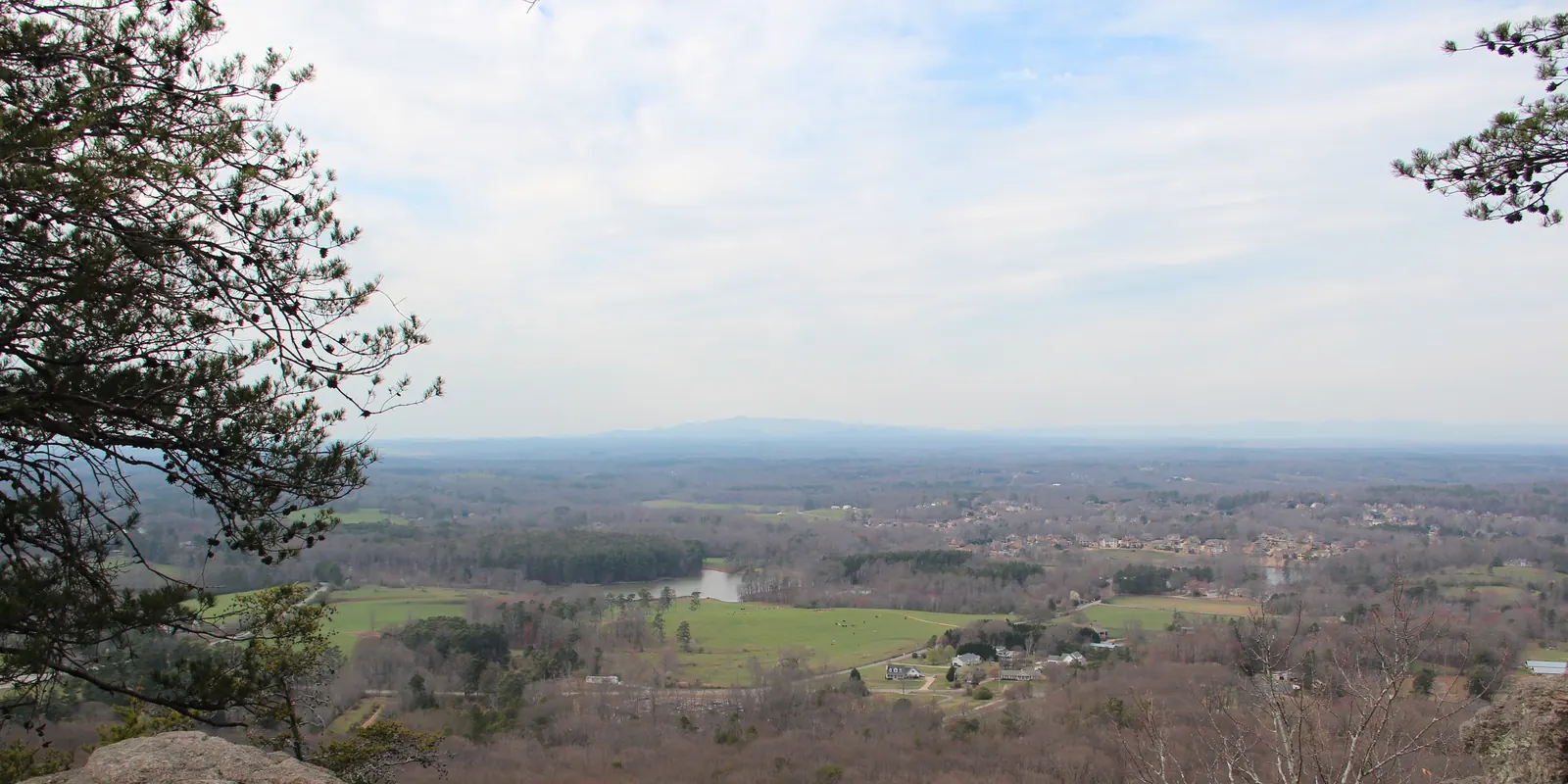 From a ledge of large granite boulders framed by a pine branch at left, a wide view over a late-winter landscape of brown woodland, green fields, small ponds and scattered subdivisions stretches to a faint line of distant mountains under a hazy white sky.