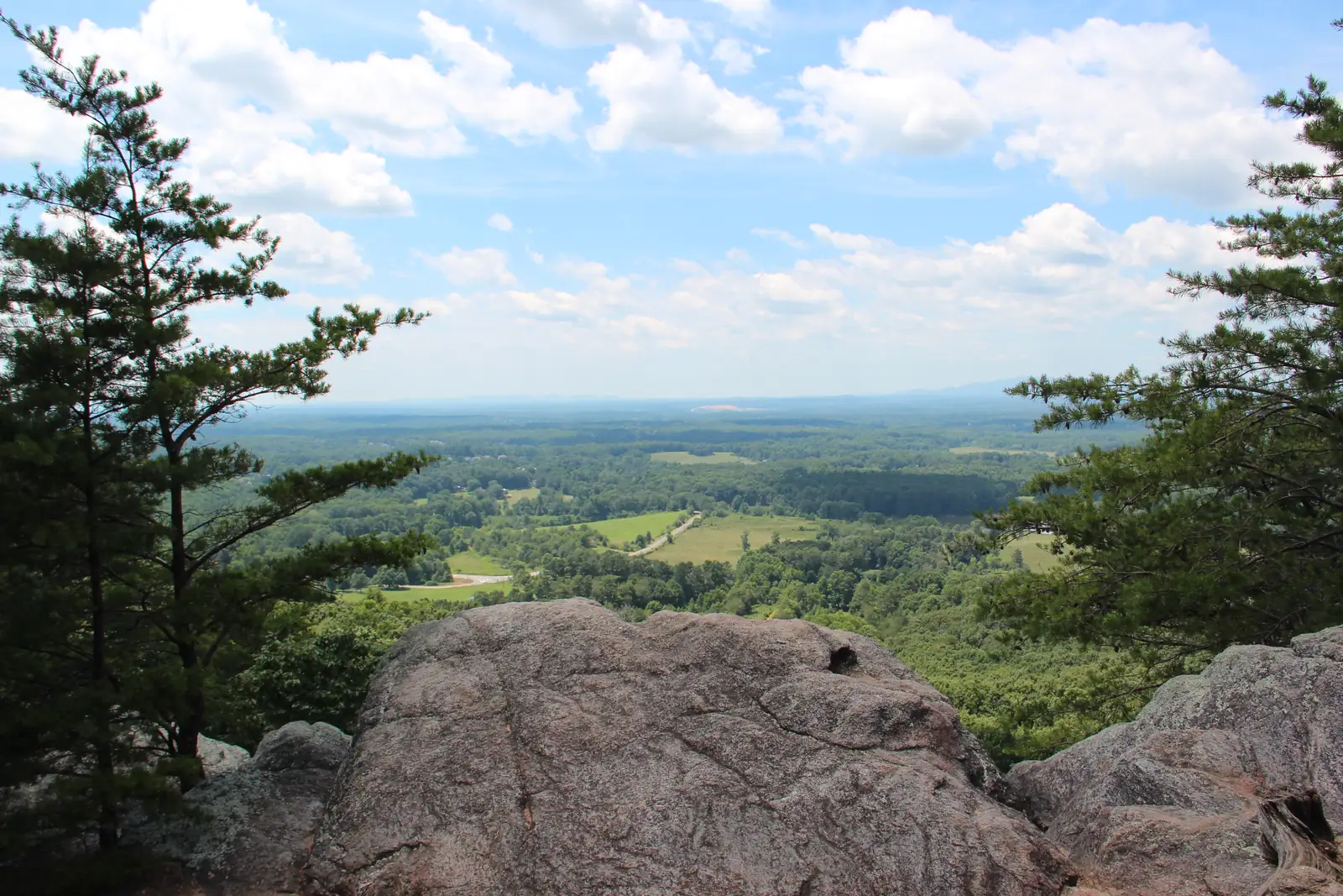 A pink-grey granite boulder outcrop in the foreground is framed by tall pines on both sides, opening onto a green valley of forest and fields beneath a bright blue sky full of white cumulus clouds.