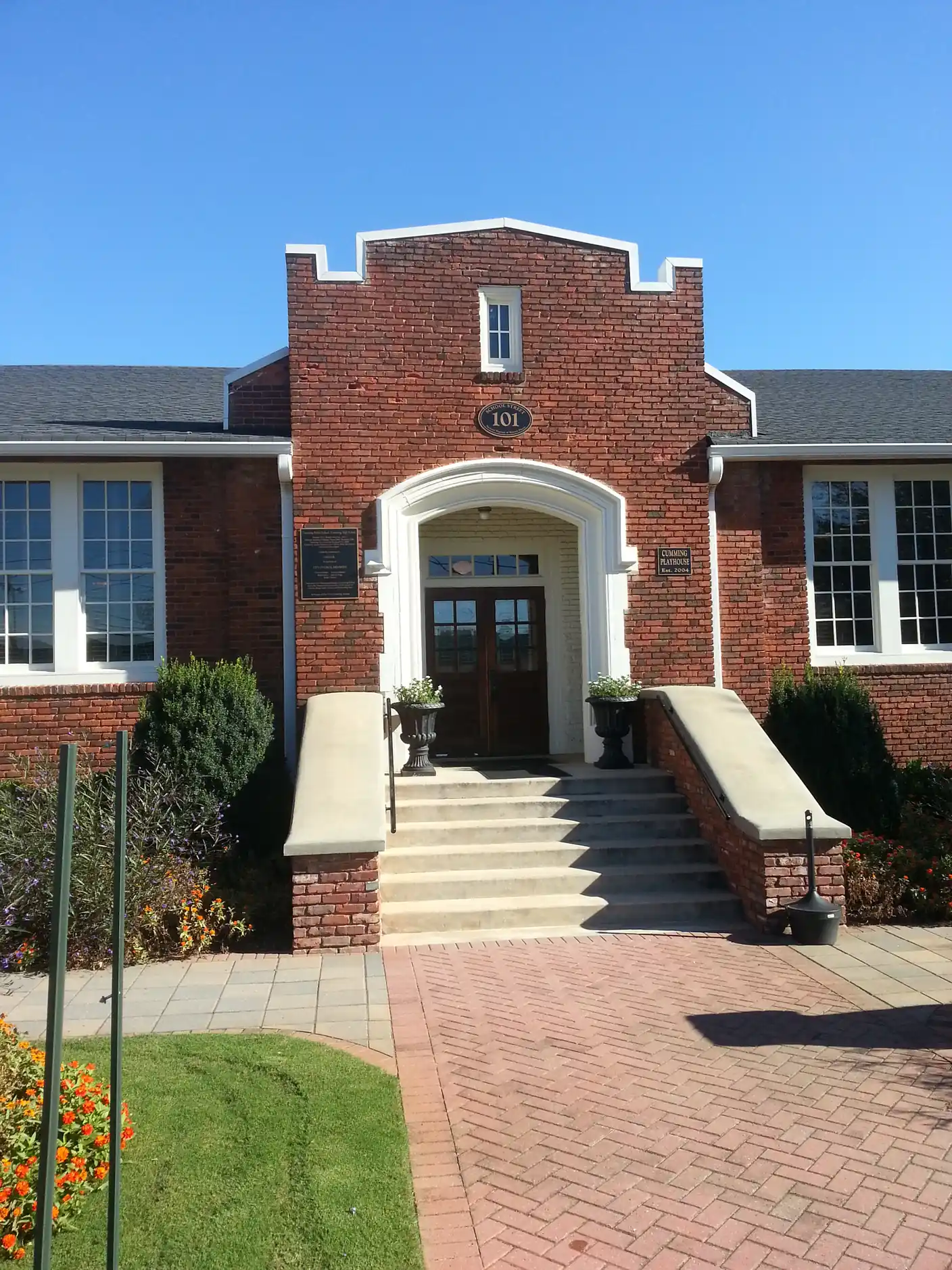 Portrait view of the entrance of a one-story red-brick building with a stepped brick parapet, a white arched doorway with double wooden doors, an oval '101' address plaque, white-framed multi-pane windows, stone-capped stair walls with urns and a herringbone brick walk lined with shrubs and orange flowers.